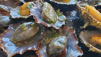 A close-up of cooked shellfish, likely clams or oysters, garnished with herbs. The shells are open, revealing the cooked meat which is shiny and moist. Some shells have yellow and green herbs visible.