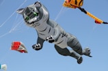 A large inflatable kite shaped like a gray cat with green eyes is flying in a clear blue sky. The cat kite appears to be mid-air with paws extended as if diving or pouncing. Other smaller kites with red and yellow patterns are also visible in the background.