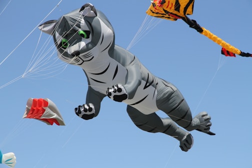 A large inflatable kite shaped like a gray cat with green eyes is flying in a clear blue sky. The cat kite appears to be mid-air with paws extended as if diving or pouncing. Other smaller kites with red and yellow patterns are also visible in the background.