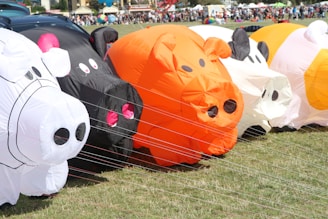 A series of inflatable animal shapes, resembling cows and pigs, are lined up on a grassy field. Brightly colored inflatables, including white, black, and orange, are tethered with strings to the ground. In the background, a crowd of people and festival tents can be seen, suggesting a festive outdoor event.