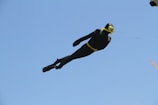 A kite designed to resemble a scuba diver is flying against a clear blue sky. The kite is mostly black with yellow accents, including goggles and a belt.