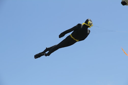 A kite designed to resemble a scuba diver is flying against a clear blue sky. The kite is mostly black with yellow accents, including goggles and a belt.