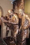 Bronze statues of three men in traditional attire, engrossed in examining a large map or document. The scene is set against a relief background depicting an urban environment with buildings. The expressions and body language suggest deep concentration and discussion.