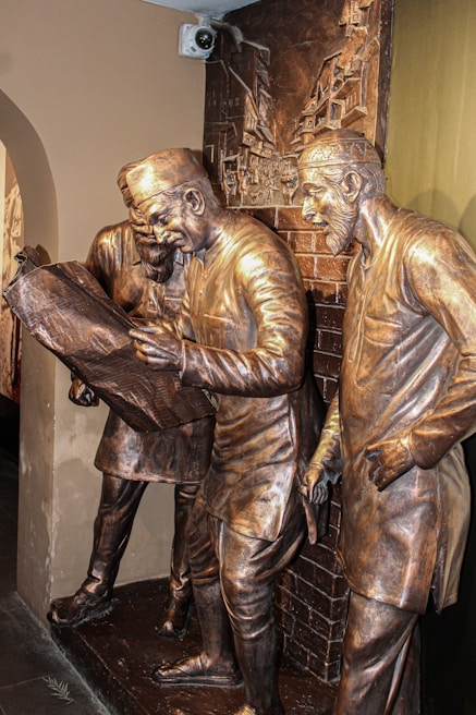 Bronze statues of three men in traditional attire, engrossed in examining a large map or document. The scene is set against a relief background depicting an urban environment with buildings. The expressions and body language suggest deep concentration and discussion.