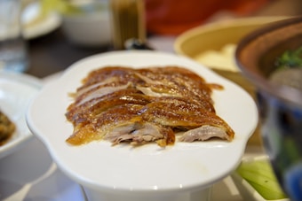 Sliced portions of roasted duck are carefully arranged on a white oval plate. The duck skin appears crispy with a glossy sheen, while the meat is tender and succulent. The background shows blurred elements of other dishes and utensils, suggesting a dining setting.