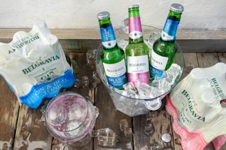 Three green bottles with colorful labels are placed in an ice-filled bucket. On either side, packages of gin and tonic bottles are visible, and a glass filled with a pink-toned drink and ice sits nearby. The setup is on a wooden surface.