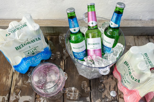 Three green bottles with colorful labels are placed in an ice-filled bucket. On either side, packages of gin and tonic bottles are visible, and a glass filled with a pink-toned drink and ice sits nearby. The setup is on a wooden surface.