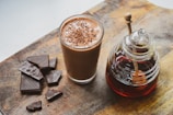 A glass filled with a chocolatey beverage topped with chocolate shavings is next to a collection of dark chocolate pieces and a jar containing a dark liquid with a honey dipper. Everything is placed on a rustic wooden surface.