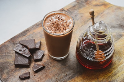A glass filled with a chocolatey beverage topped with chocolate shavings is next to a collection of dark chocolate pieces and a jar containing a dark liquid with a honey dipper. Everything is placed on a rustic wooden surface.