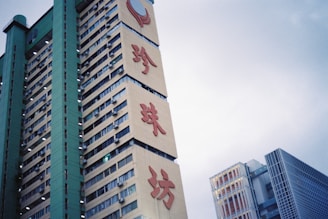 A tall building with a green and beige facade featuring large red Chinese characters prominently displayed. Several windows line the structure, some with air conditioning units installed. A modern glass building is visible in the background, set against a cloudy sky.