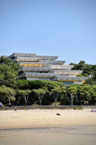 A modern, multi-level building with large balconies and an unusual layered design, surrounded by lush greenery. The structure is located near a sandy beach, with a promenade separating the building from the shore. People can be seen relaxing on the beach and walking in the area.