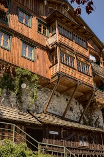 A rustic building constructed with wood and stone features green-framed windows and ornate wooden balconies. The structure is adorned with climbing plants and decorative elements, such as antlers hanging on the wall. Sunlight illuminates the facade, highlighting the aged texture of the materials.
