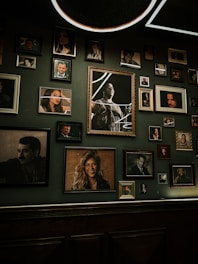 A collection of framed portraits hanging on a dimly lit wall. The frames vary in size and style, displaying images of various individuals, some in black and white and others in color. The wall appears to have a dark green hue, giving the space an intimate and vintage ambiance.
