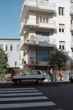 A multi-story residential building with balconies featuring a white exterior. Two people walk past a parked small white car on a street near a pedestrian crossing. The building has closed shutters and decorative items on balconies. Trees and a fence line the sidewalk.