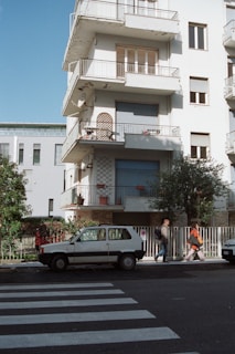A multi-story residential building with balconies featuring a white exterior. Two people walk past a parked small white car on a street near a pedestrian crossing. The building has closed shutters and decorative items on balconies. Trees and a fence line the sidewalk.