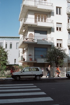 A multi-story residential building with balconies featuring a white exterior. Two people walk past a parked small white car on a street near a pedestrian crossing. The building has closed shutters and decorative items on balconies. Trees and a fence line the sidewalk.