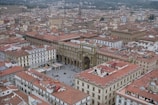 Aerial view of an expansive cityscape featuring historical buildings with red-tiled roofs. A prominent stone archway stands at the center of a bustling square, surrounded by pedestrian activity and a few parked cars. The dense cluster of buildings extends into the distance, with a mix of architectural styles visible.