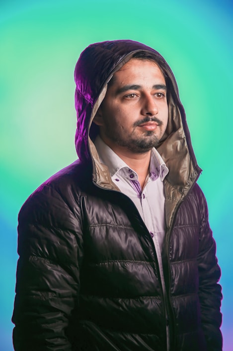 A man wearing a hooded, dark puffer jacket stands against a vibrant, gradient background of teal and blue. His expression is neutral, with soft lighting highlighting his facial features.