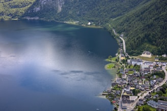 Aerial view of a peaceful lakeside town with a large expanse of calm water reflecting the sky. The town is nestled at the foot of lush green mountains, with a mix of residential buildings and roads lining the shore. A winding road runs through the landscape, connecting the community amidst the natural scenery.