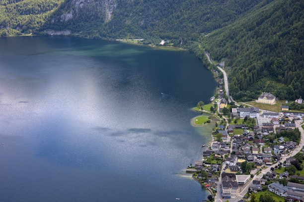 Aerial view of a peaceful lakeside town with a large expanse of calm water reflecting the sky. The town is nestled at the foot of lush green mountains, with a mix of residential buildings and roads lining the shore. A winding road runs through the landscape, connecting the community amidst the natural scenery.