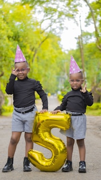 Two children wearing party hats and matching outfits stand outdoors on a tree-lined path. They hold a large, shiny gold balloon shaped like the number five. Both are smiling and appear to be celebrating.