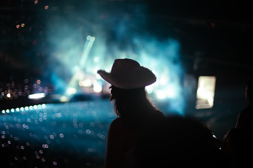 A silhouette of a person wearing a cowboy hat is seen in the foreground, facing a concert stage with bright lights and smoke effects. The scene is filled with bokeh, creating a dreamy effect with lights scattered throughout the background.