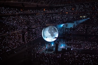 A large concert audience fills a stadium, illuminated by glowing hand-held lights. A massive, realistic moon projection dominates the center stage, casting a mystical ambiance over the crowd. Stage lights and equipment are visible around the moon.