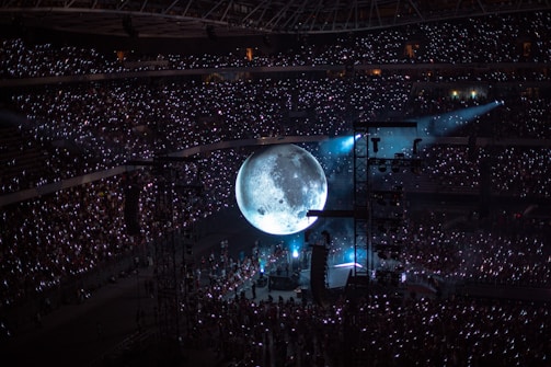 A large concert audience fills a stadium, illuminated by glowing hand-held lights. A massive, realistic moon projection dominates the center stage, casting a mystical ambiance over the crowd. Stage lights and equipment are visible around the moon.