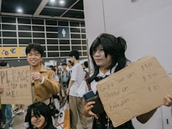 A group of people at a convention, possibly cosplaying, with one individual holding a cardboard sign that reads 'PLEASE STEP ON ME!' Another person next to them holds a sign offering fees for actions like being 'sat on,' 'stepped on,' and 'kabedon'. They are surrounded by a crowd of attendees in a large, indoor space with signage and overhead lighting visible.