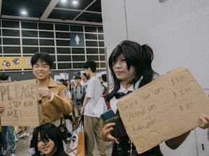 A group of people at a convention, possibly cosplaying, with one individual holding a cardboard sign that reads 'PLEASE STEP ON ME!' Another person next to them holds a sign offering fees for actions like being 'sat on,' 'stepped on,' and 'kabedon'. They are surrounded by a crowd of attendees in a large, indoor space with signage and overhead lighting visible.