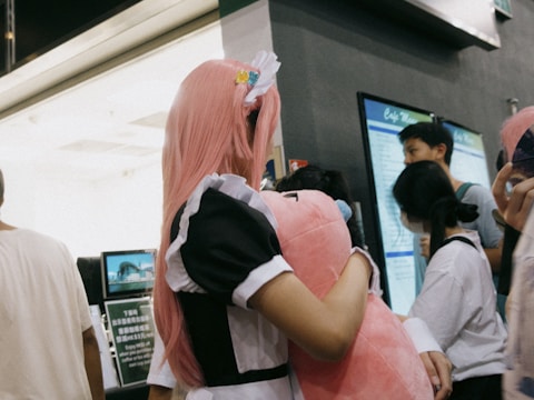 A person with long pink hair is wearing a black and white outfit resembling a maid costume. They are holding a large pink plush toy. In the background, there are groups of people gathered, some looking at a display board or advertisement.