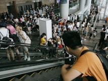A crowded scene at a convention or public event with numerous people on escalators and the main floor. Many attendees appear to be dressed in various costumes, suggesting a cosplay or anime-related event. The environment is bustling, with people interacting and moving in different directions.