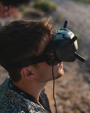 A person with short brown hair is wearing black virtual reality goggles with antennas, looking upwards. The setting appears to be outdoors on a sunny day, with a blurry background of what may be grass or a path.