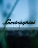A large sign with the word 'Lamborghini' in bold letters is mounted on top of a building. The foreground appears slightly blurred, possibly due to foliage framing the sign. The background is a clear blue sky.