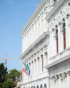 A grand, ornate building with classical architecture featuring a series of columns and decorative stonework. Italian and European Union flags are attached to the building. In the background, a construction crane is visible next to some trees under a clear blue sky.