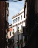 A narrow street flanked by tall, old buildings with European architectural features. The buildings have windows adorned with small balconies and green plants. A sign for 'Tabacchi' is visible, indicating a tobacco shop. The sky visible above is clear and blue.