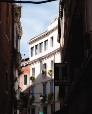 A narrow street flanked by tall, old buildings with European architectural features. The buildings have windows adorned with small balconies and green plants. A sign for 'Tabacchi' is visible, indicating a tobacco shop. The sky visible above is clear and blue.
