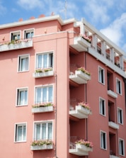 A residential building painted in a warm pink color, featuring multiple floors with balconies. Each balcony is adorned with flower boxes filled with blooming flowers. The building is set against a bright blue sky with a few white clouds, and there is a metal railing on the balconies.