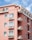 A residential building painted in a warm pink color, featuring multiple floors with balconies. Each balcony is adorned with flower boxes filled with blooming flowers. The building is set against a bright blue sky with a few white clouds, and there is a metal railing on the balconies.