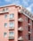 A residential building painted in a warm pink color, featuring multiple floors with balconies. Each balcony is adorned with flower boxes filled with blooming flowers. The building is set against a bright blue sky with a few white clouds, and there is a metal railing on the balconies.