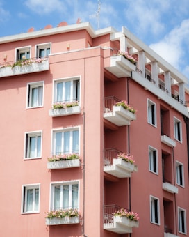 A residential building painted in a warm pink color, featuring multiple floors with balconies. Each balcony is adorned with flower boxes filled with blooming flowers. The building is set against a bright blue sky with a few white clouds, and there is a metal railing on the balconies.