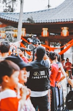A group of people in colorful traditional clothing gathered, with a focus on a camera operator filming the event. The background features a pavilion with lanterns and decorative red ribbons.