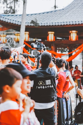 A group of people in colorful traditional clothing gathered, with a focus on a camera operator filming the event. The background features a pavilion with lanterns and decorative red ribbons.