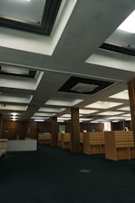 A spacious room with a dark carpeted floor and an arrangement of wooden cabinets that appear to be card catalog files. The ceiling features a grid of rectangular recessed lighting panels and air conditioning vents. The walls are adorned with a couple of framed artworks.