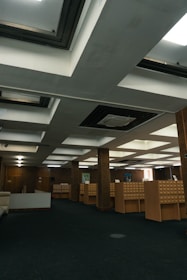 A spacious room with a dark carpeted floor and an arrangement of wooden cabinets that appear to be card catalog files. The ceiling features a grid of rectangular recessed lighting panels and air conditioning vents. The walls are adorned with a couple of framed artworks.