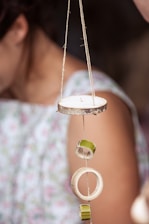 A string with circular wooden pieces and green rings is suspended vertically. In the background, part of a person wearing a floral-patterned dress is visible.