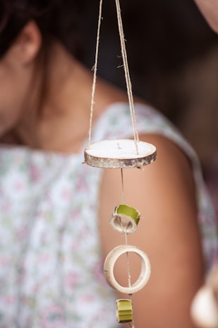 A string with circular wooden pieces and green rings is suspended vertically. In the background, part of a person wearing a floral-patterned dress is visible.