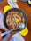 A dish featuring several large prawns cooked in a skillet, garnished with lemon slices and herbs. The prawns are arranged neatly and accompanied by a spoon resting on the edge of the skillet. The setting includes a wooden table, a napkin, fork, and knife, adding to the dining ambiance.