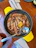 A dish featuring several large prawns cooked in a skillet, garnished with lemon slices and herbs. The prawns are arranged neatly and accompanied by a spoon resting on the edge of the skillet. The setting includes a wooden table, a napkin, fork, and knife, adding to the dining ambiance.