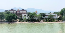 A peaceful riverside scene with a large multi-story building on the left and smaller white structures on the right nestled among lush green trees. The river in the foreground has a calm surface and mountains appear hazy in the background under a clear sky.